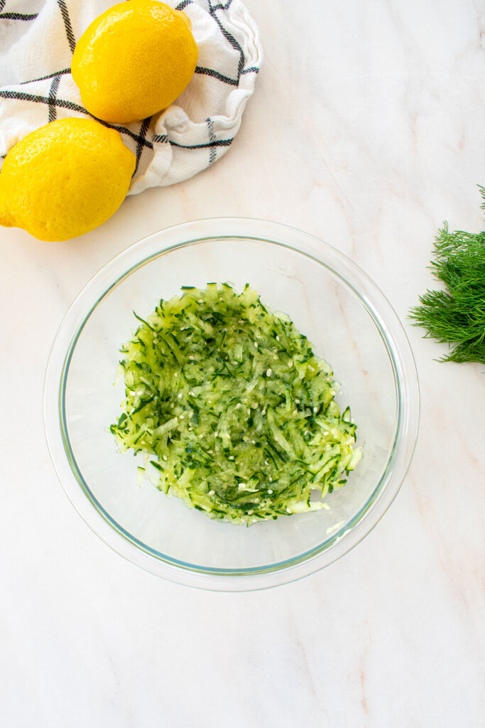 grated cucumber in a glass mixing bowl on a marble countertop.