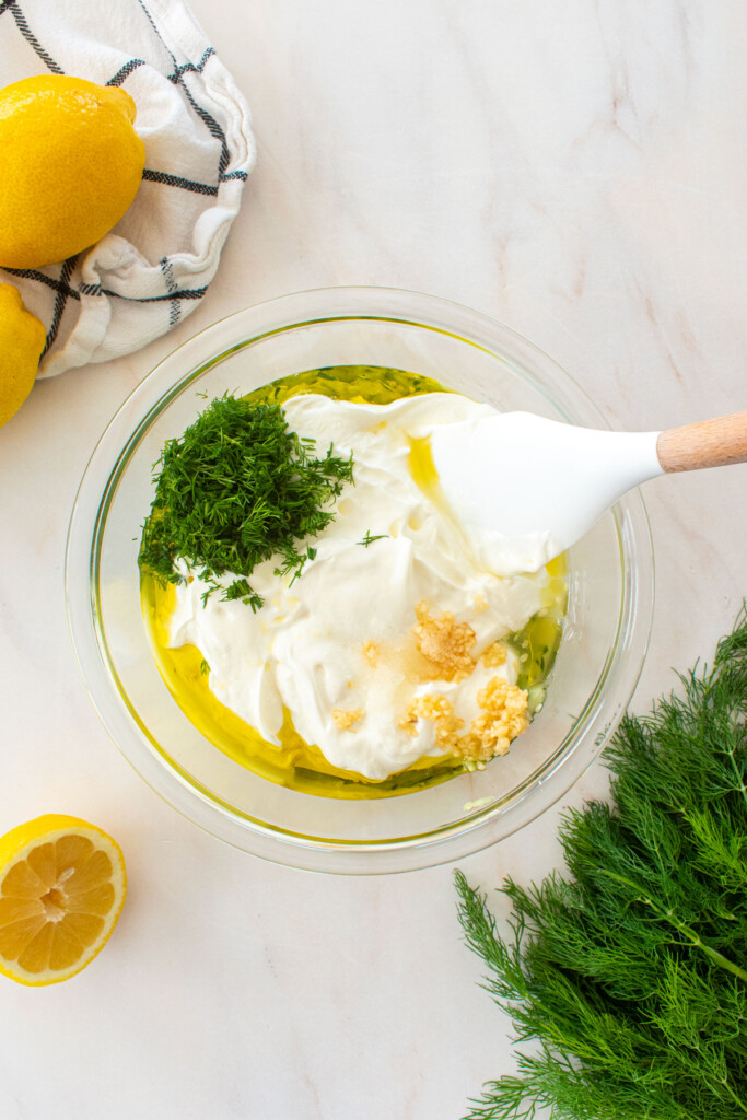 ingredients for organic tzatziki sauce with fresh dill in a glass mixing bowl on a marble countertop.