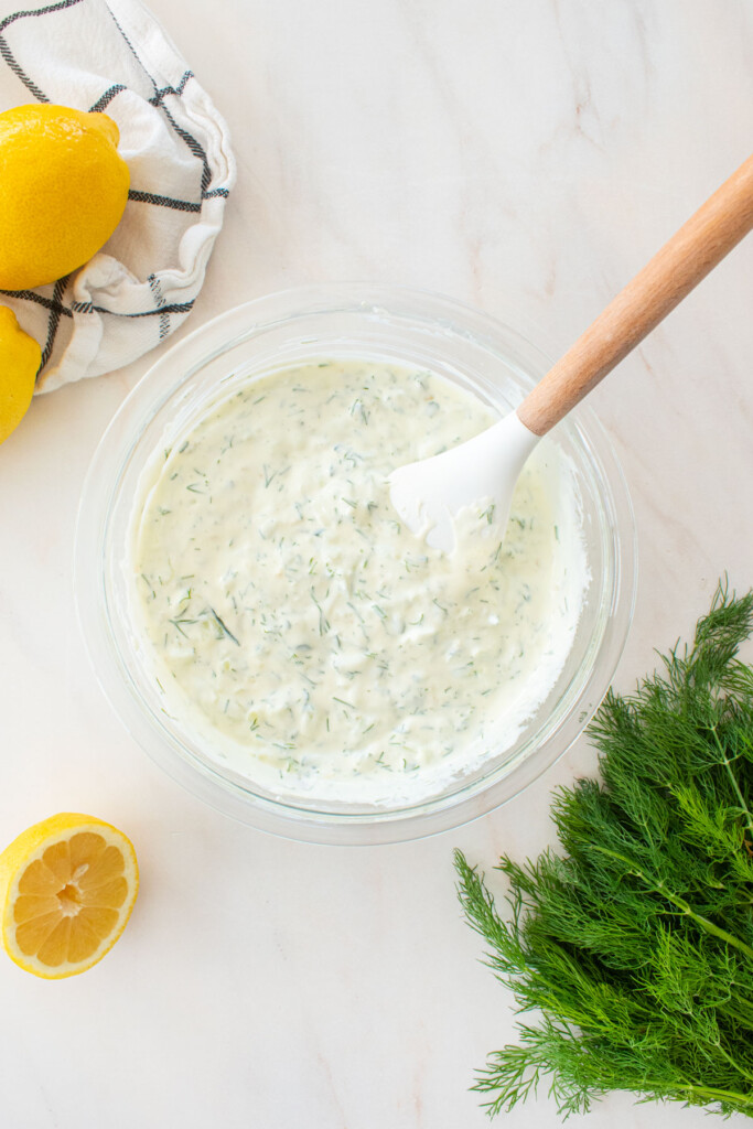 organic tzatziki sauce with fresh dill in a glass mixing bowl on a marble countertop.