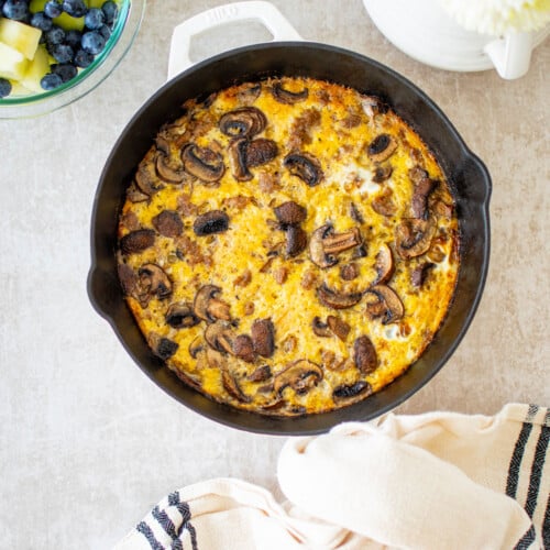 bone broth sausage mushroom frittata in a cast iron skillet on the table with a bowl of fruit.