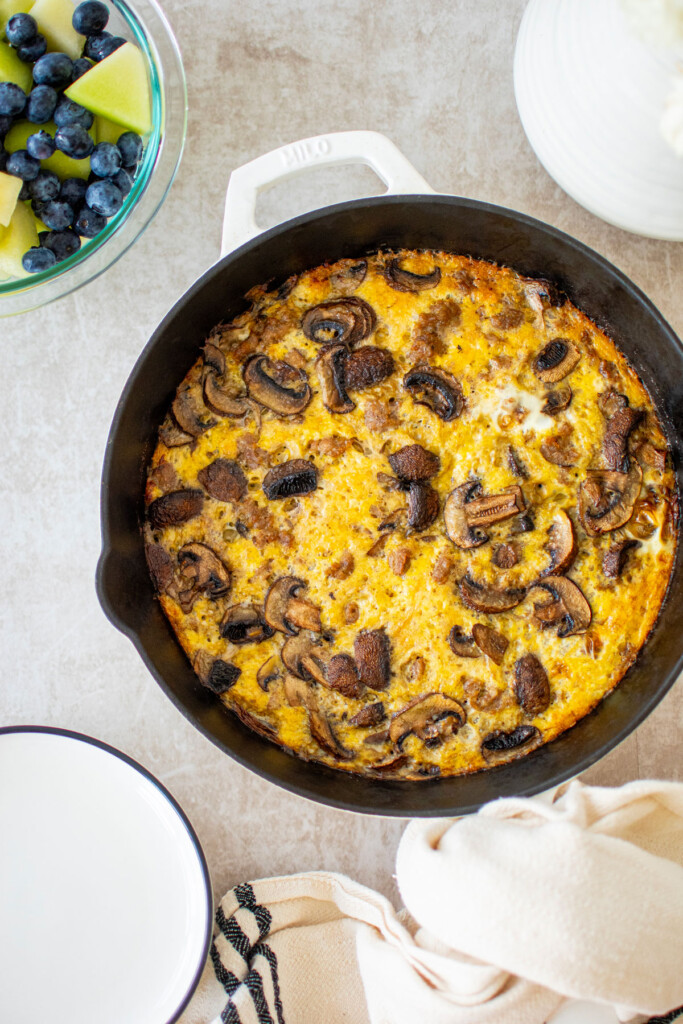 bone broth sausage mushroom frittata in a cast iron skillet on the table with a bowl of fruit.