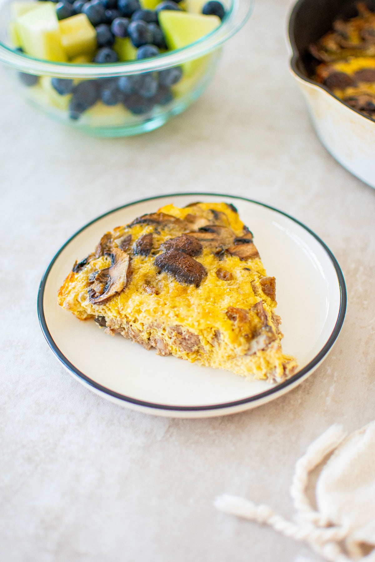 slice of bone broth sausage mushroom frittata on a plate on the breakfast table with a bowl of fruit.