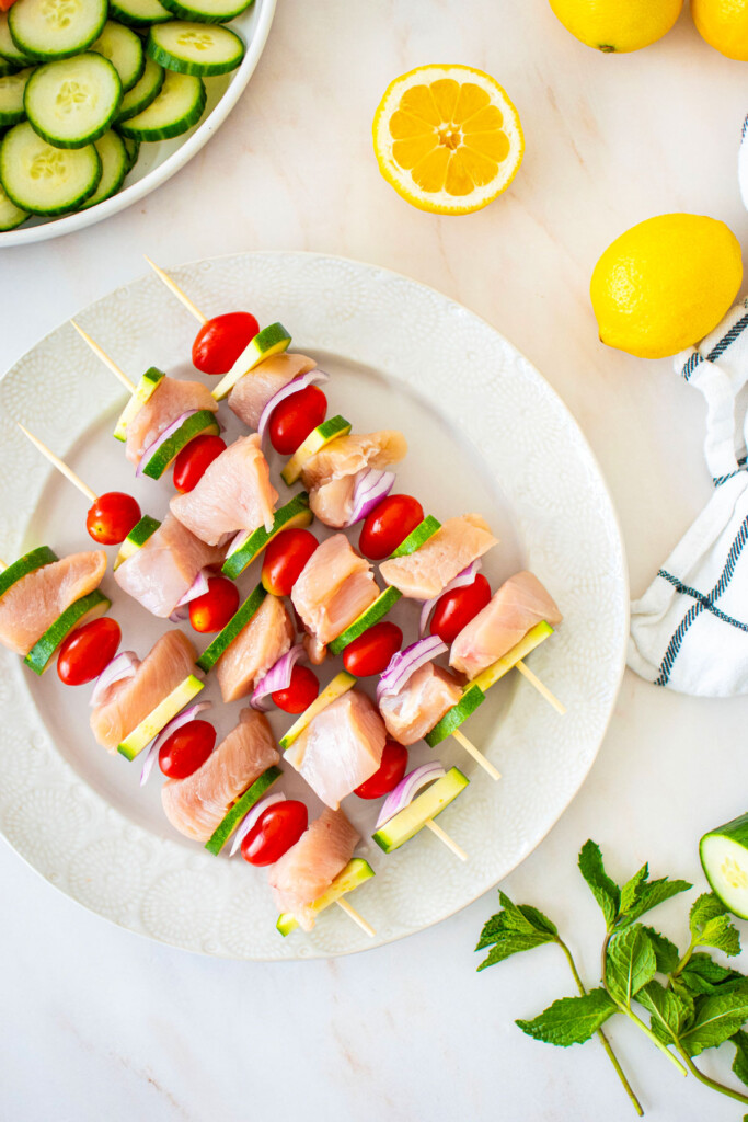 raw chicken, zucchini, red onion, tomato on skewers on a white plate on a marble countertop.