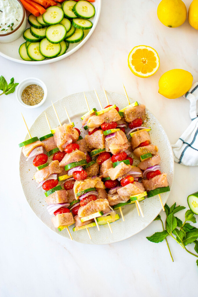 raw chicken, zucchini, red onion, tomato and spices on skewers on a white plate on a marble countertop.