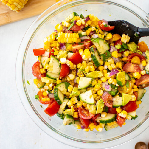 real simple corn salad with avocado being mixed in a glass mixing bowl on a white countertop.