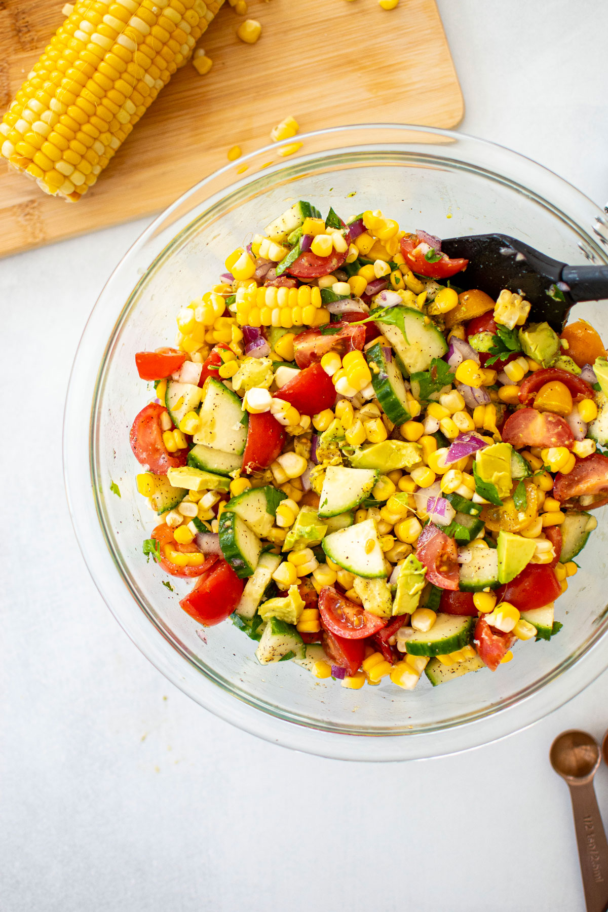 real simple corn salad with avocado being mixed in a glass mixing bowl on a white countertop.