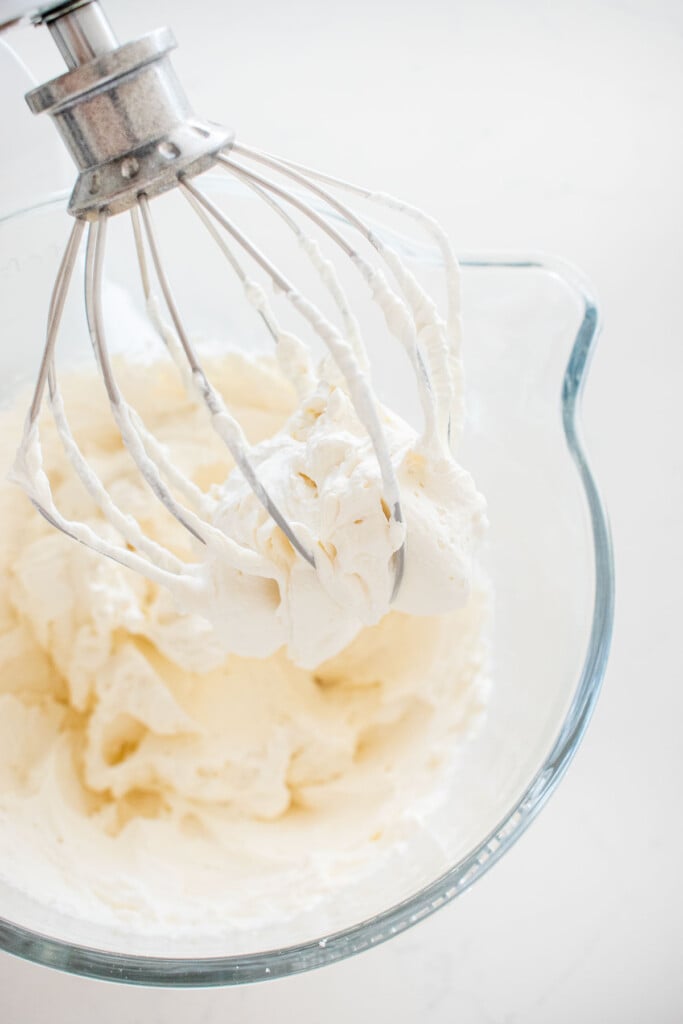 homemade whipped cream frosting in a stand mixer on a white marble counter.