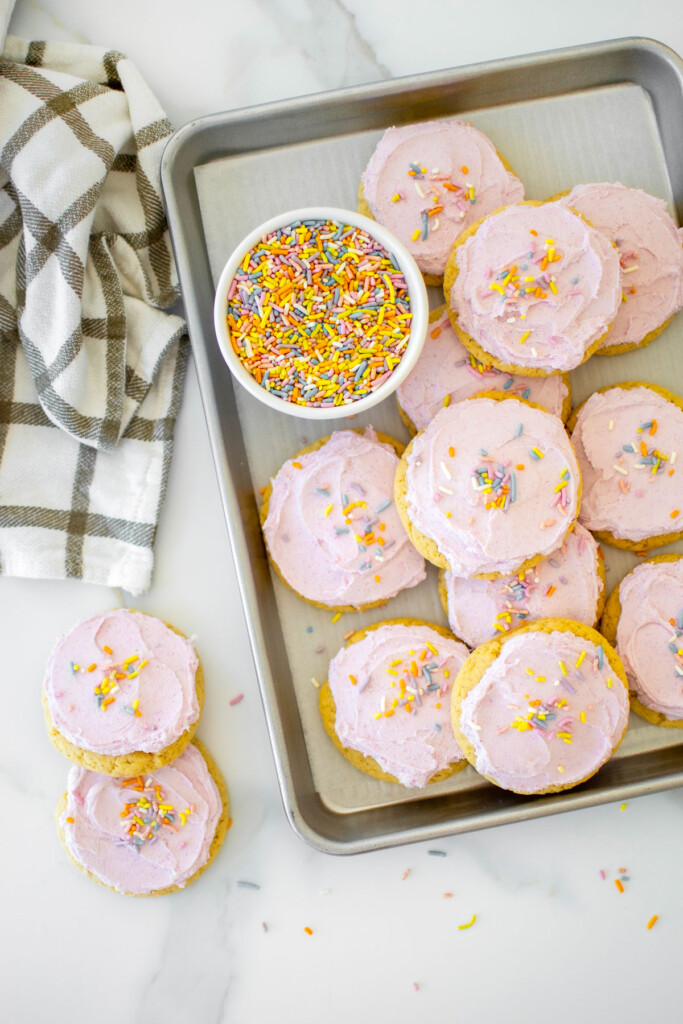 soft sugar cookies with pink frosting and rainbow sprinkles on a baking sheet on a white marble counter.