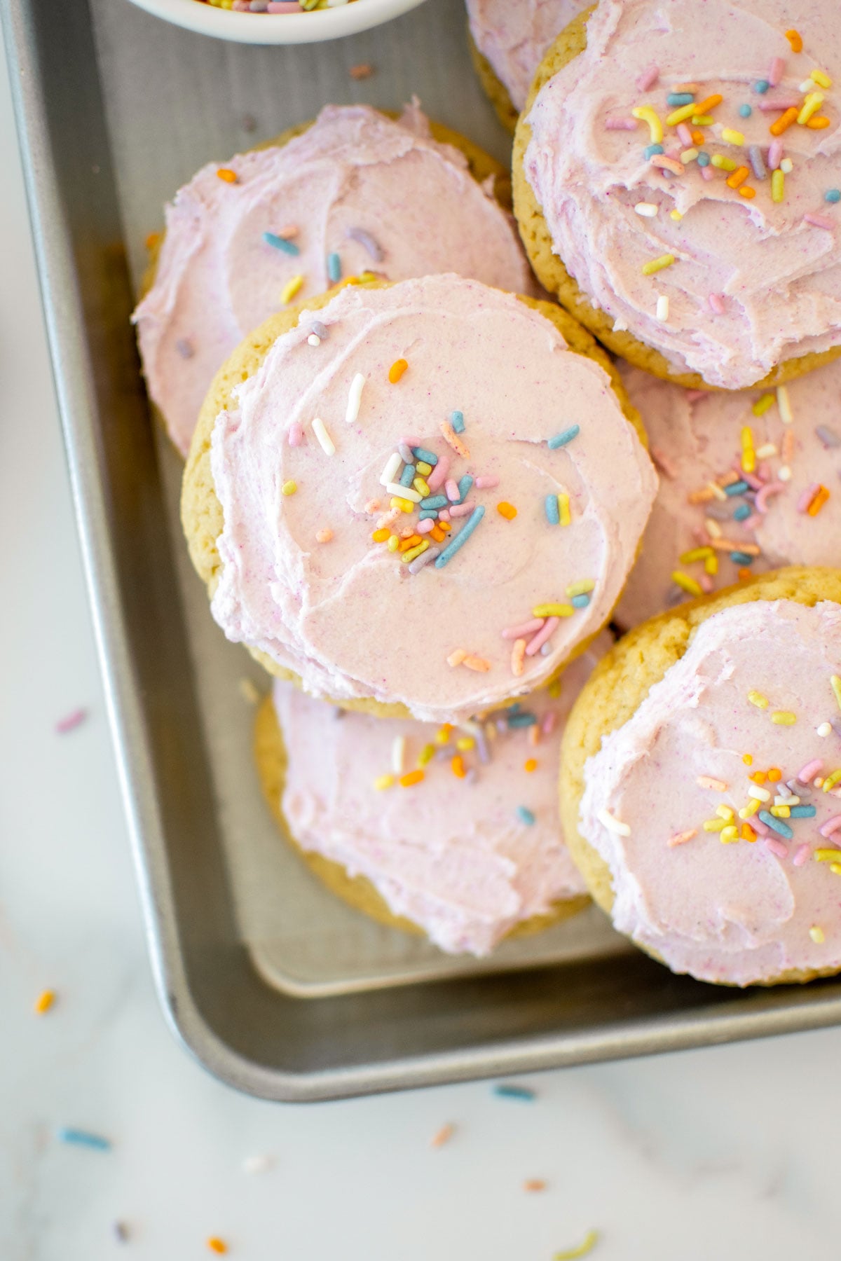 soft sugar cookies with pink frosting and rainbow sprinkles on a baking sheet on a white marble counter.