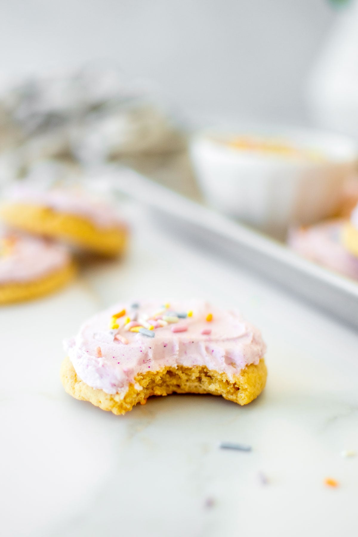soft sugar cookie with pink frosting and rainbow sprinkles with a bite taken out on a white marble counter.