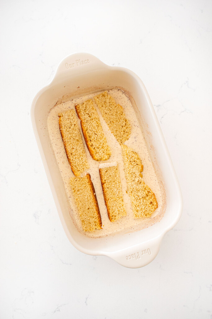 sourdough sticks of bread soaking in french toast custard batter in a shallow baking dish on a marble counter.