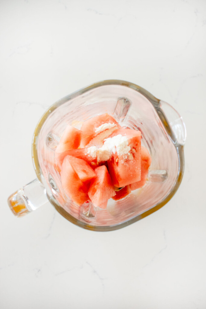 ingredients for strawberry watermelon smoothie with collagen in a glass blender on a white marble counter.