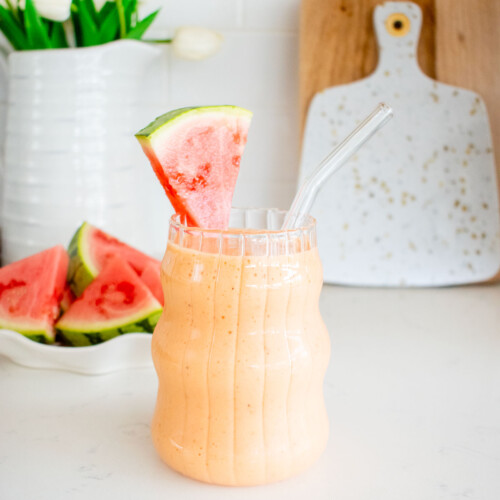 strawberry watermelon smoothie with collagen in a glass with a watermelon slice and straw on kitchen counter.