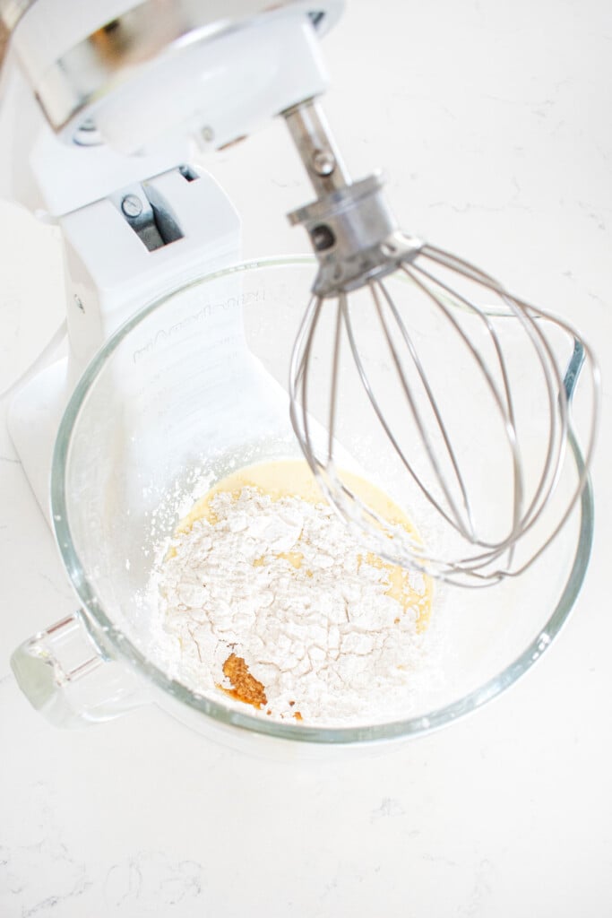 ingredients for homemade whipped cream in a stand mixing bowl with a whisk attachment on a marble counter.
