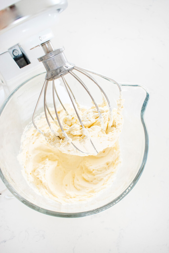 homemade whipped cream on the whisk of a glass mixing bowl on a stand mixer on a white marble counter.