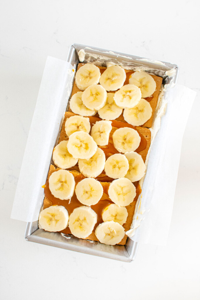 banoffee icebox cake being assembled in a loaf pan on a white marble counter.