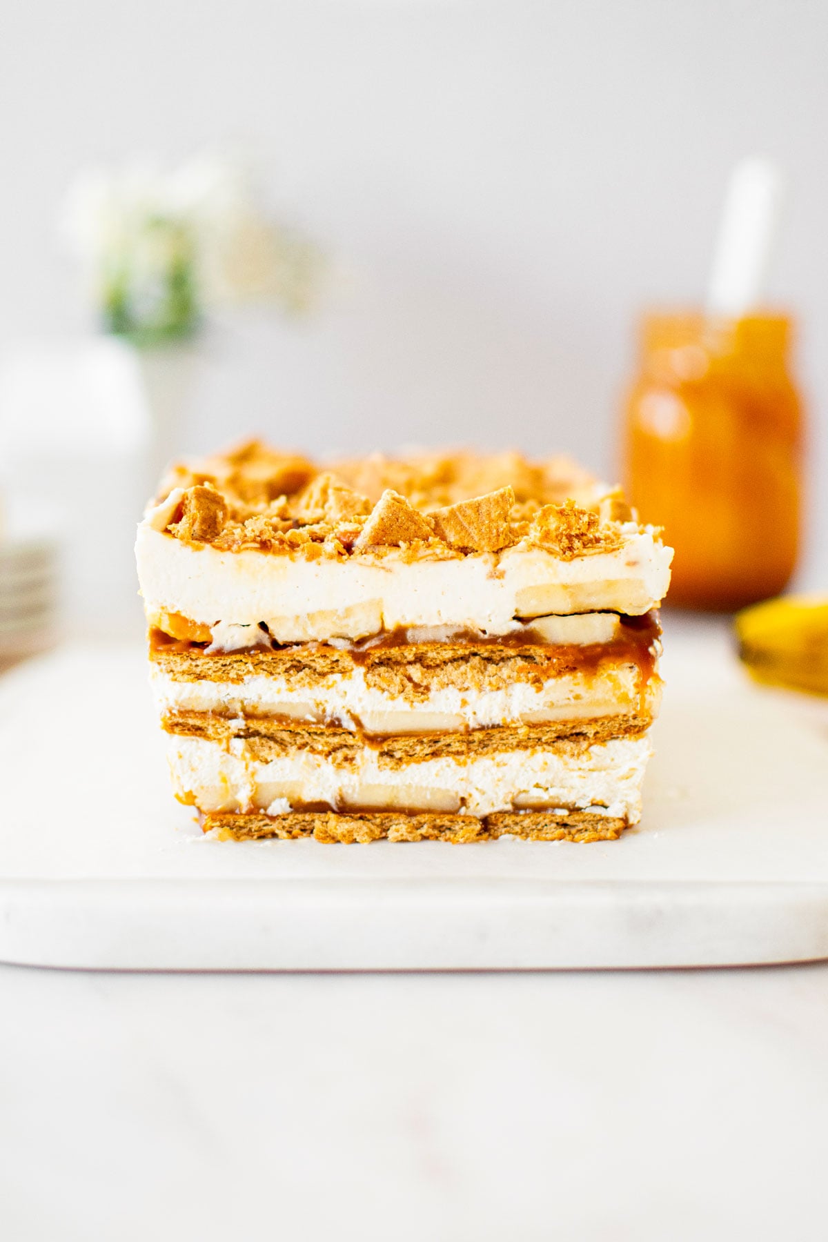 banoffee icebox cake on a white marble serving board on a table with a jar of caramel sauce and flowers.