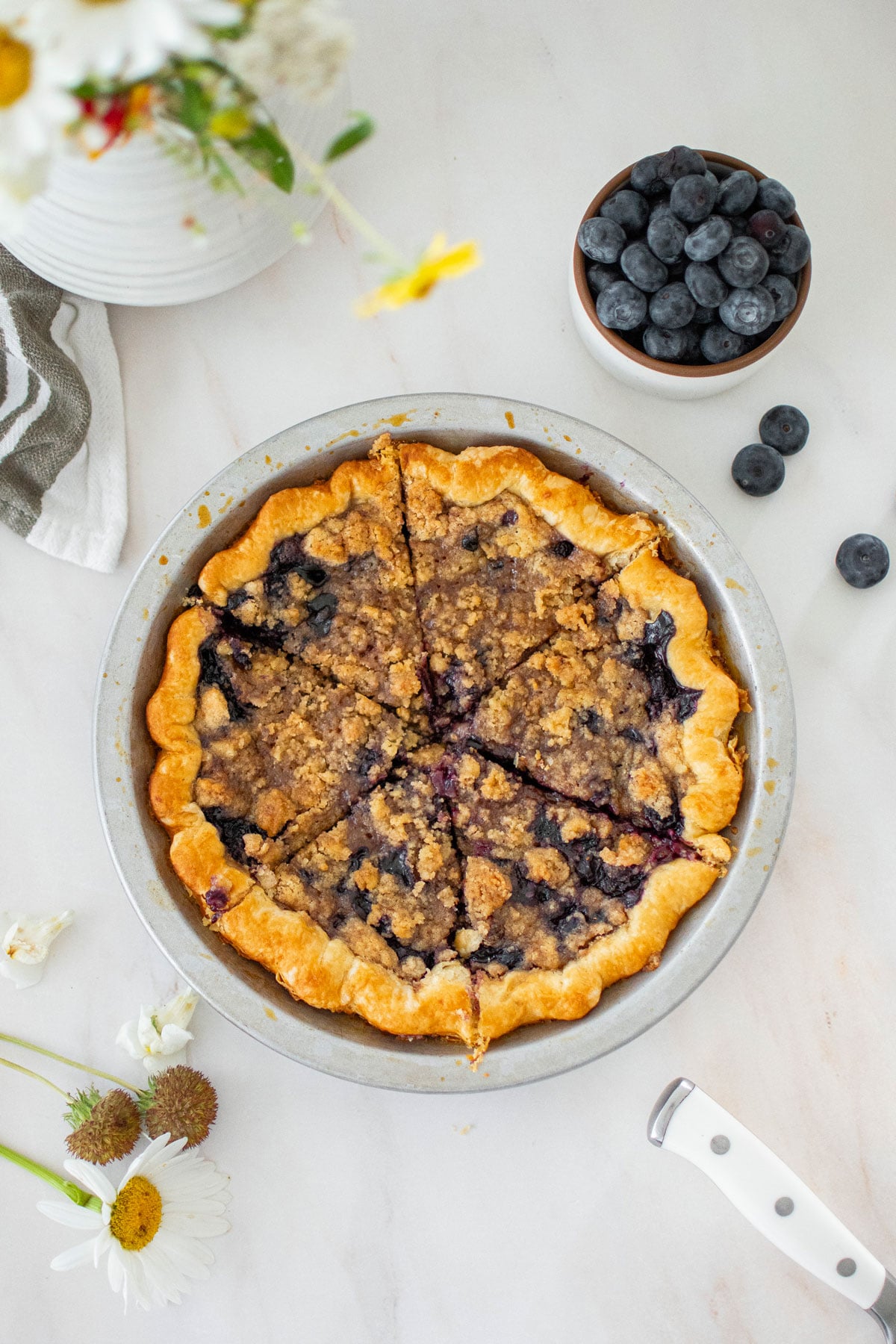 blueberry honey crumble pie in a silver pie pan sliced into slices on a marble counter.