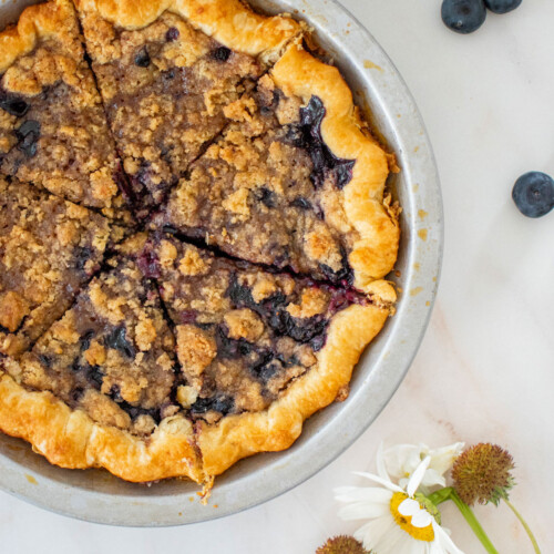 blueberry honey crumble pie in a silver pie pan sliced into slices on a marble counter.