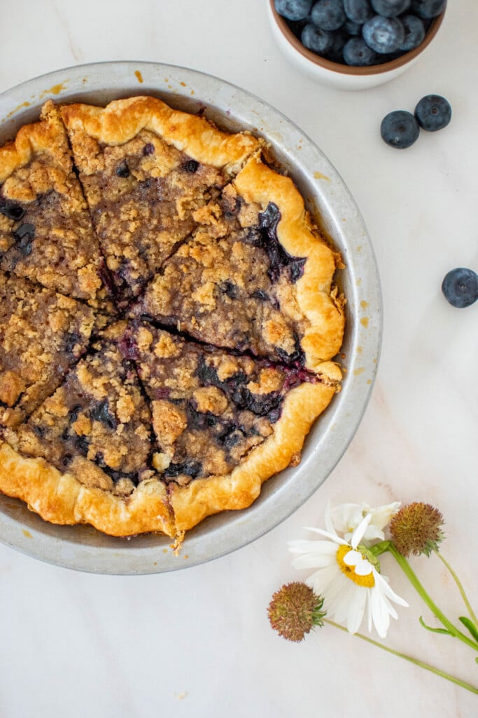 blueberry honey crumble pie in a silver pie pan sliced into slices on a marble counter.