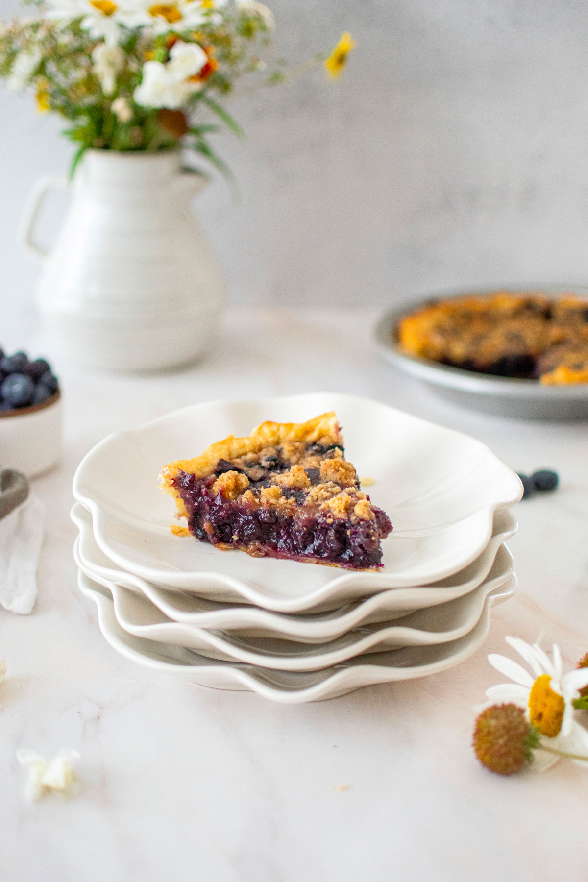 slice of blueberry honey crumble pie on a stack of white ruffle plates on a table with fresh flowers.