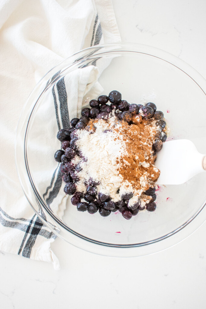 glass mixing bowl with blueberries, sugar, honey, flour, cornstarch and cinnamon on a marble counter.