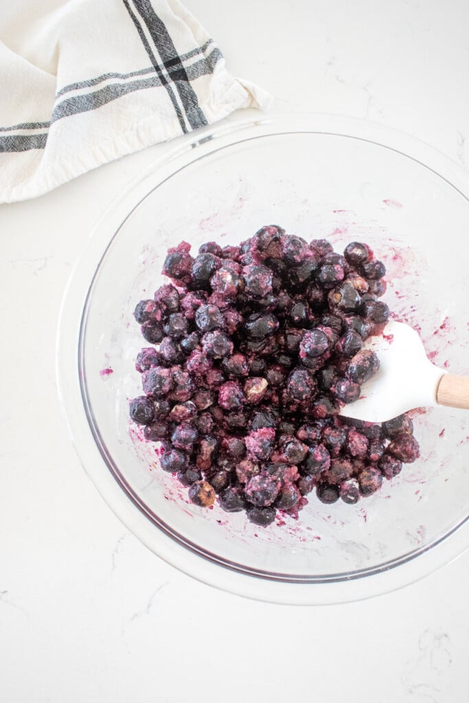 glass mixing bowl with blueberry pie filling mixed together with a spatula on a marble counter.