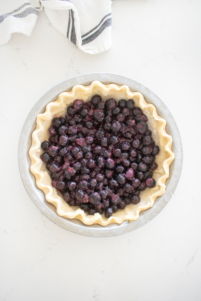 fluted pie crust with blueberry filling in an aluminum pie pan on a marble counter.