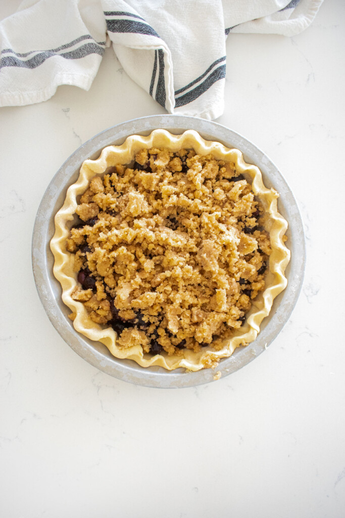 fluted pie crust with blueberry filling and crumble topping on top in an aluminum pie pan on a marble counter.