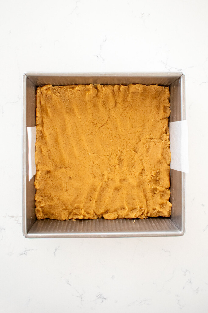 dough pressed into a square baking pan on a white marble counter.