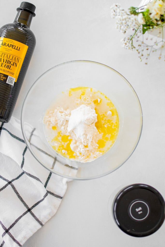 ingredients for flatbread pizza dough in a glass mixing bowl on a white table with a bottle of olive oil.
