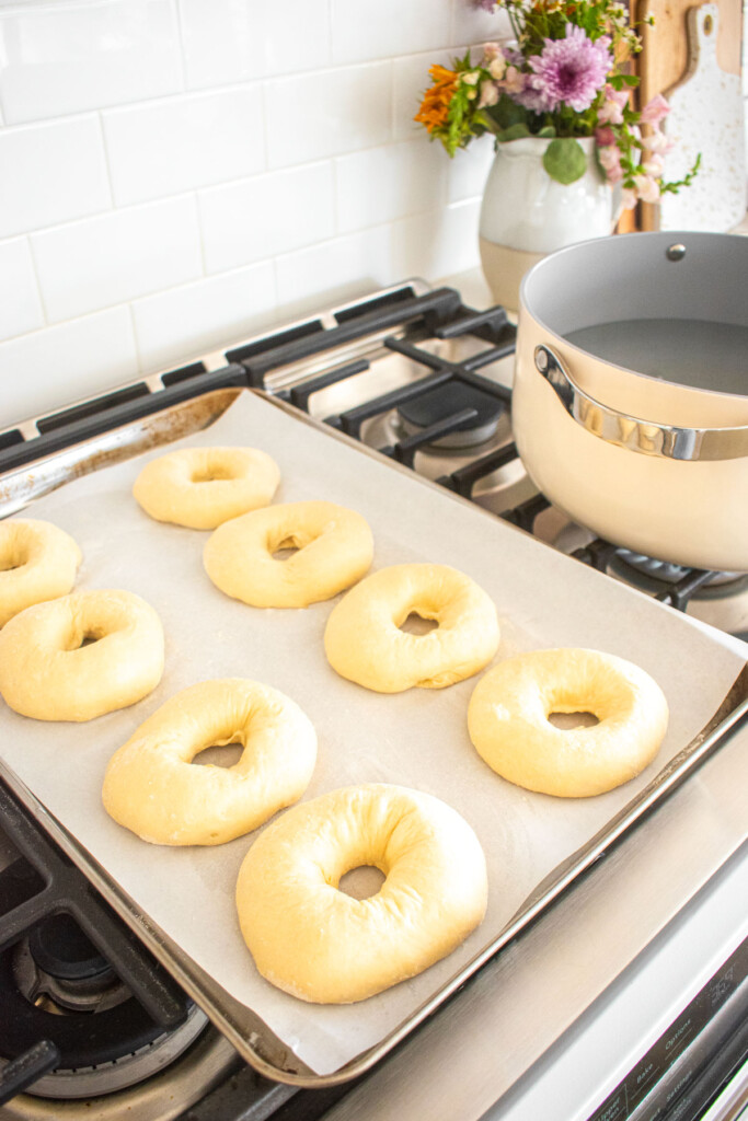homemade sesame bagel dough with holes formed on a baking sheet sitting on top of a range.