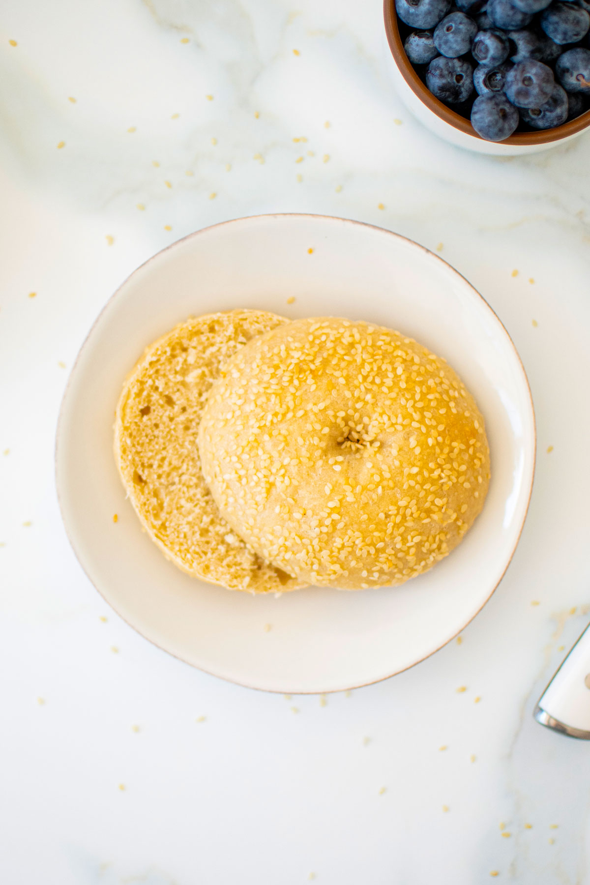 homemade sesame bagel cut in half on a white plate on a marble counter with fresh blueberries.