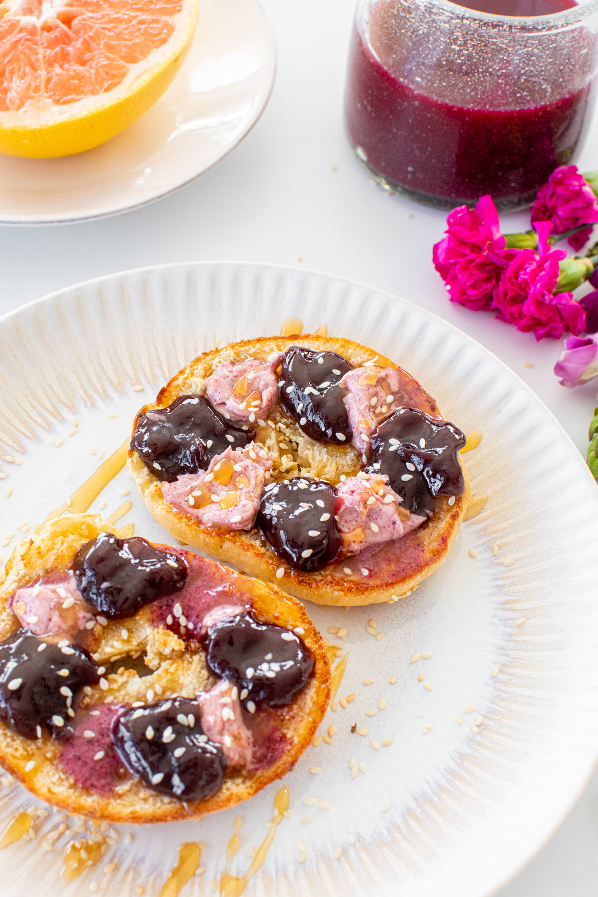 toasted sesame bagel with blackberry butter and blackberry jam with a drizzle of honey and sesame seeds on a plate on a breakfast table.