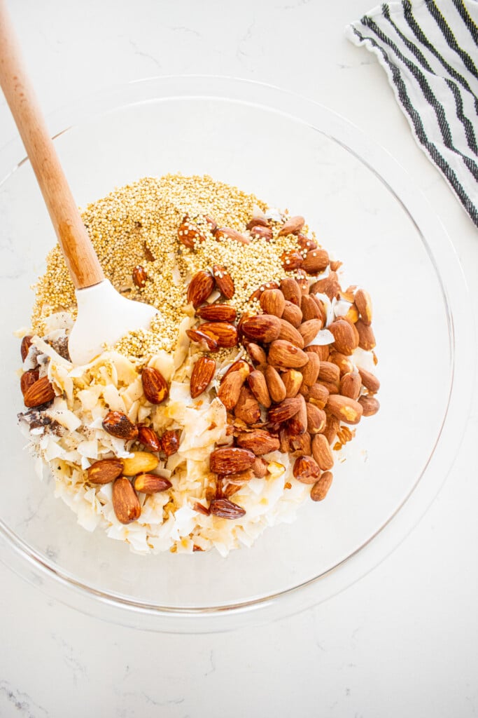 wet ingredients added into dry ingredients for honey almond coconut granola in a large glass mixing bowl with a spatula on a marble counter.