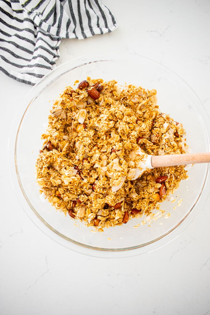 honey almond coconut granola being stirred with a spatula in a glass mixing bowl on a white marble counter.