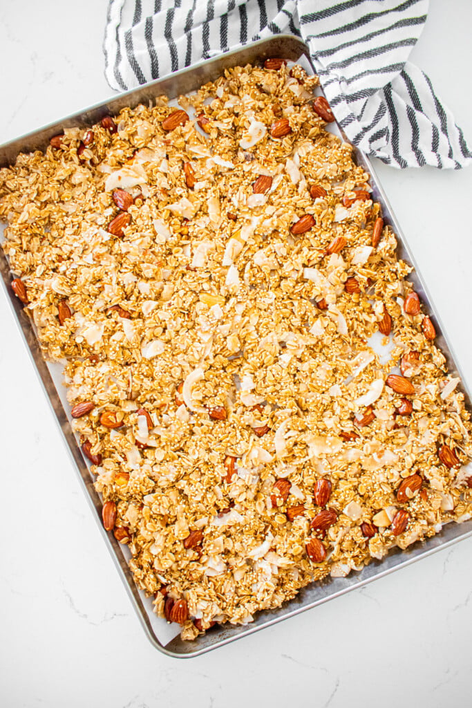 honey almond coconut granola spread out onto a baking sheet on a white marble counter