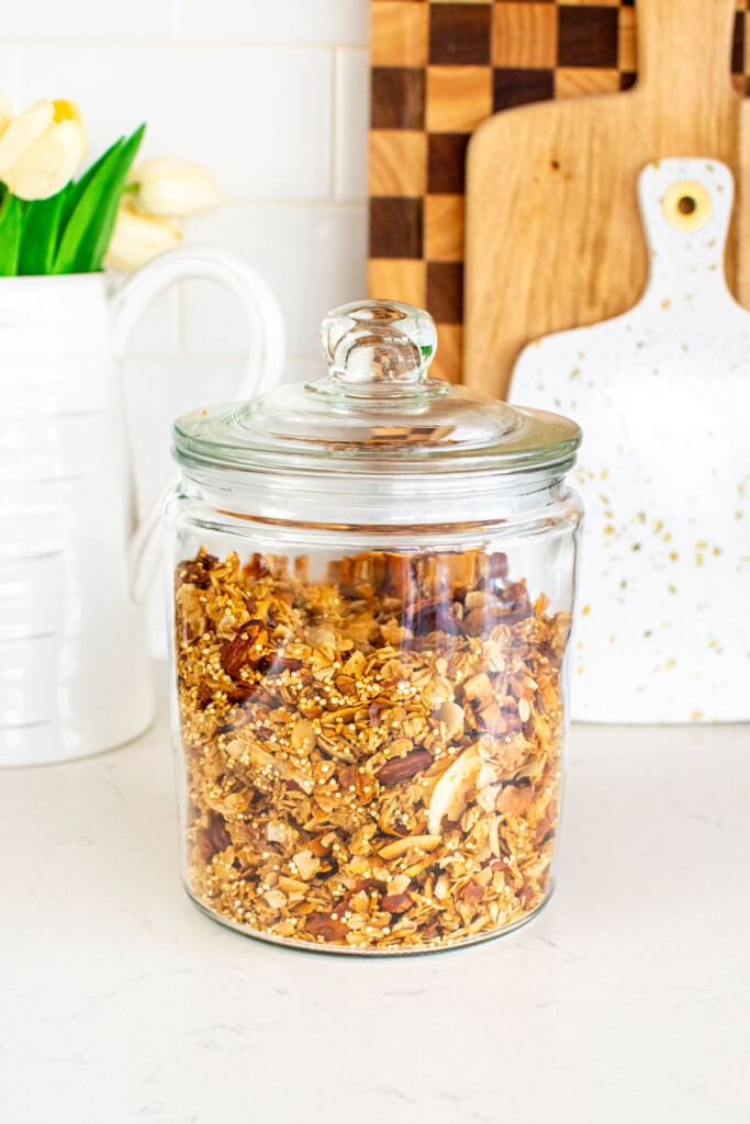 honey almond coconut granola in a glass canister on a white marble counter.