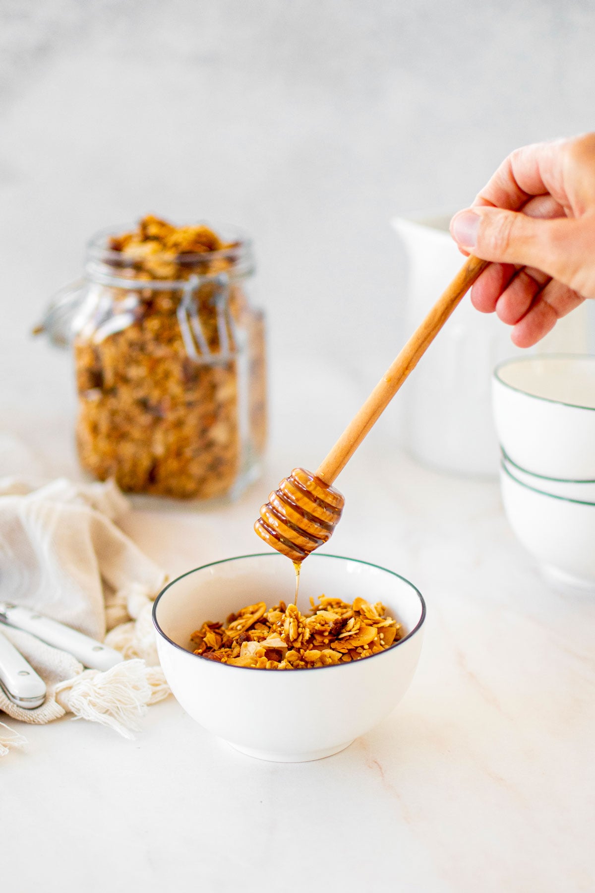 honey almond coconut granola in a bowl with a woman adding honey with a honey dipstick.