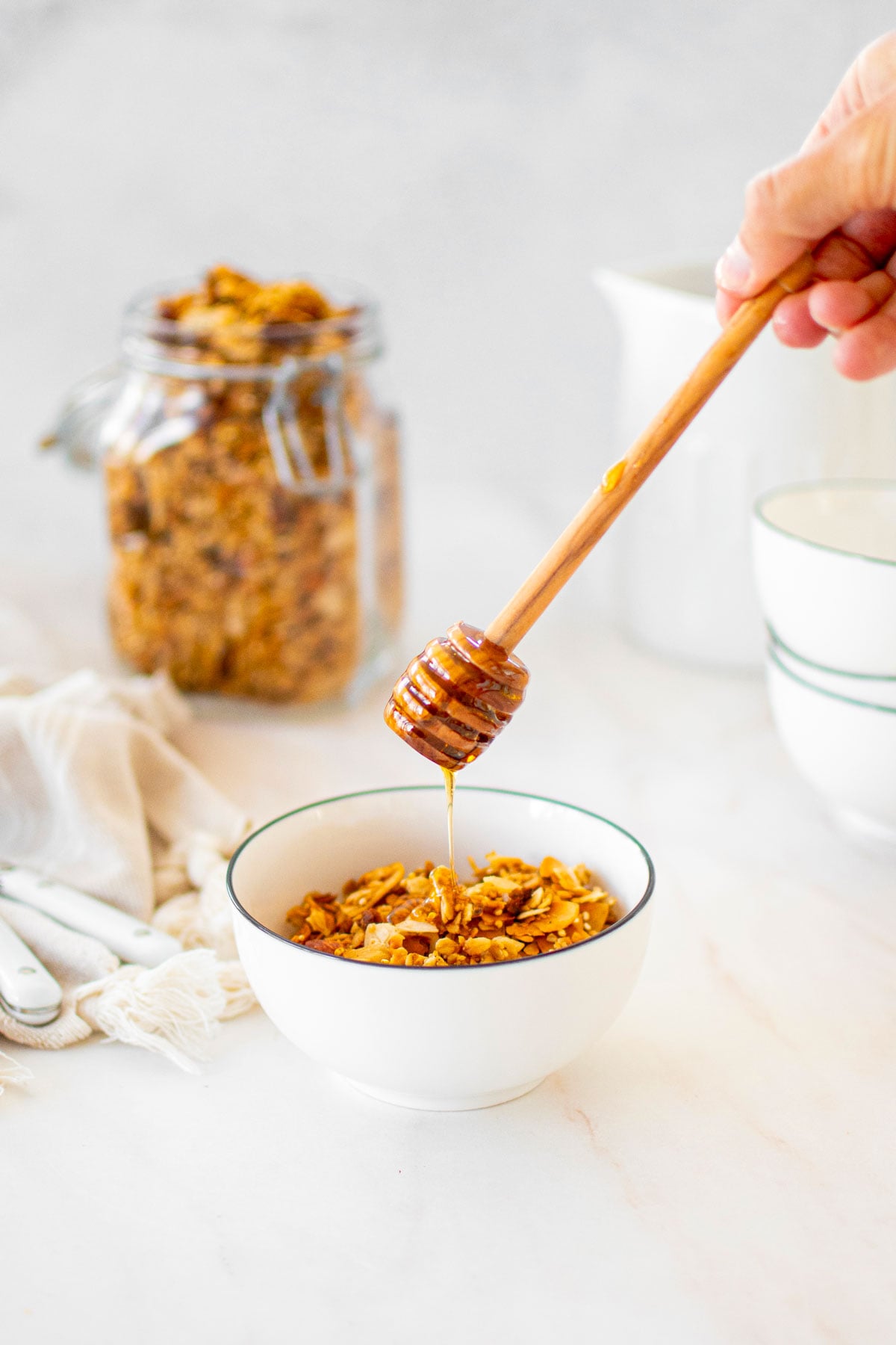 honey almond coconut granola in a bowl with a woman adding honey with a honey dipstick.