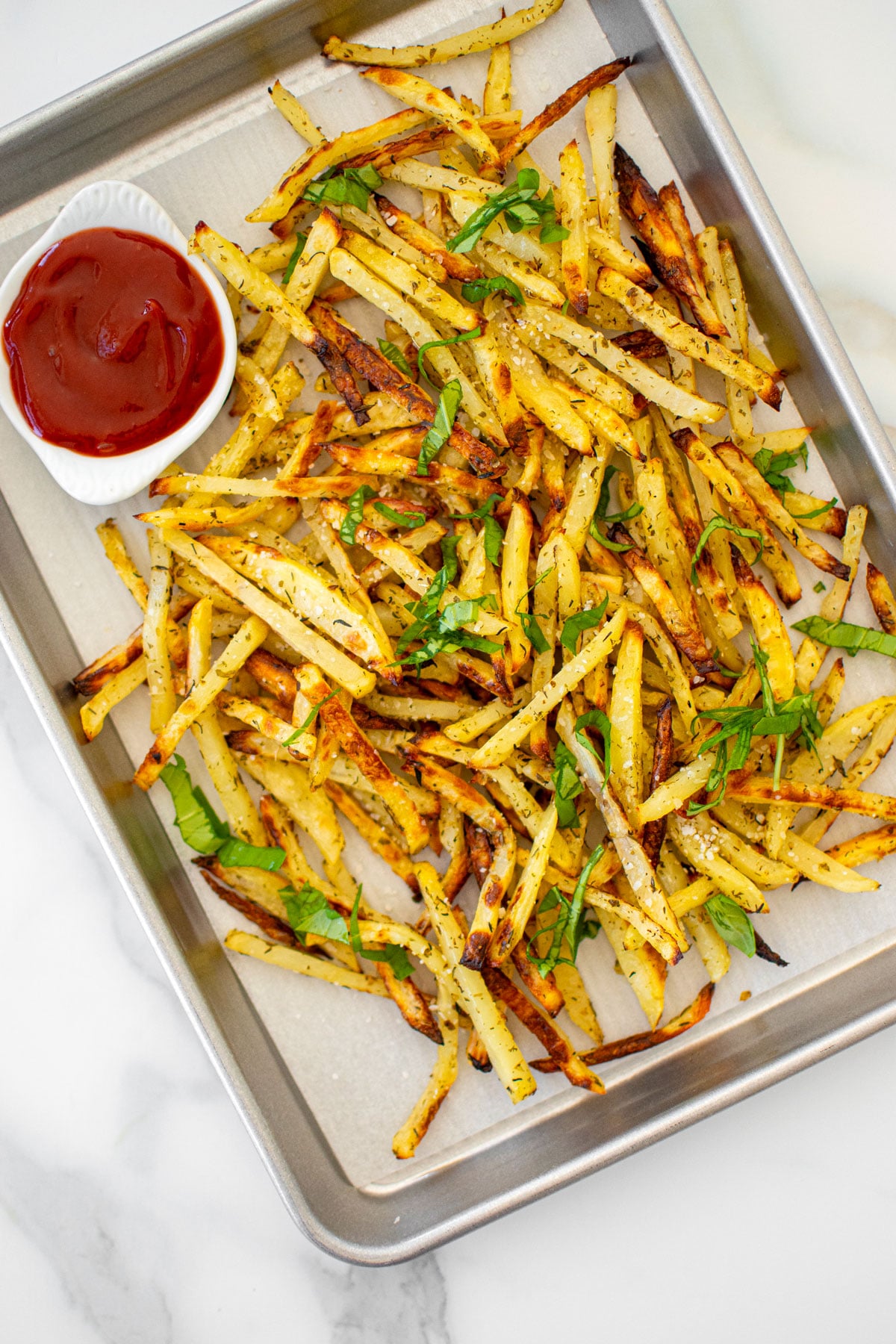 lemon herb french fries with a small dish of ketchup on a baking sheet on a white marble countertop.