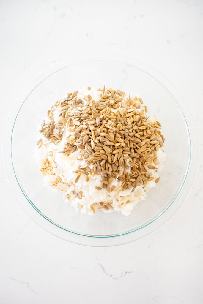 oats, coconut and sunflower seeds in a glass mixing bowl on a white marble counter.