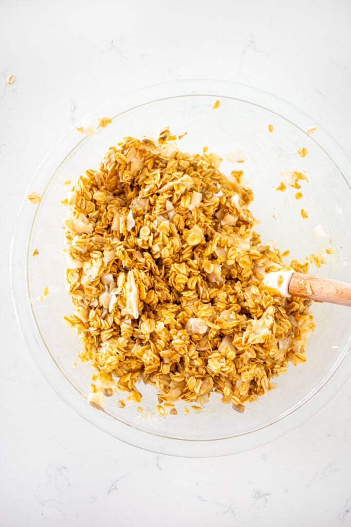 peanut butter honey granola for dogs being mixed in a glass mixing bowl on a white marble counter.
