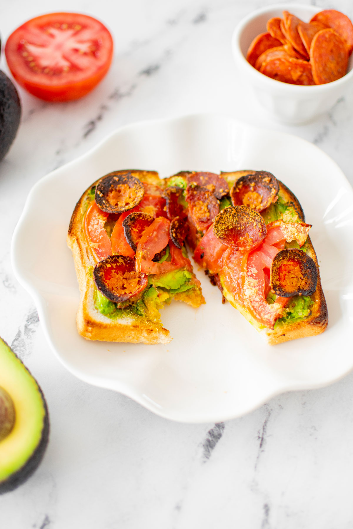 pizza avocado toast cut in half with a bite taken out of one half on a white plate on a marble countertop.