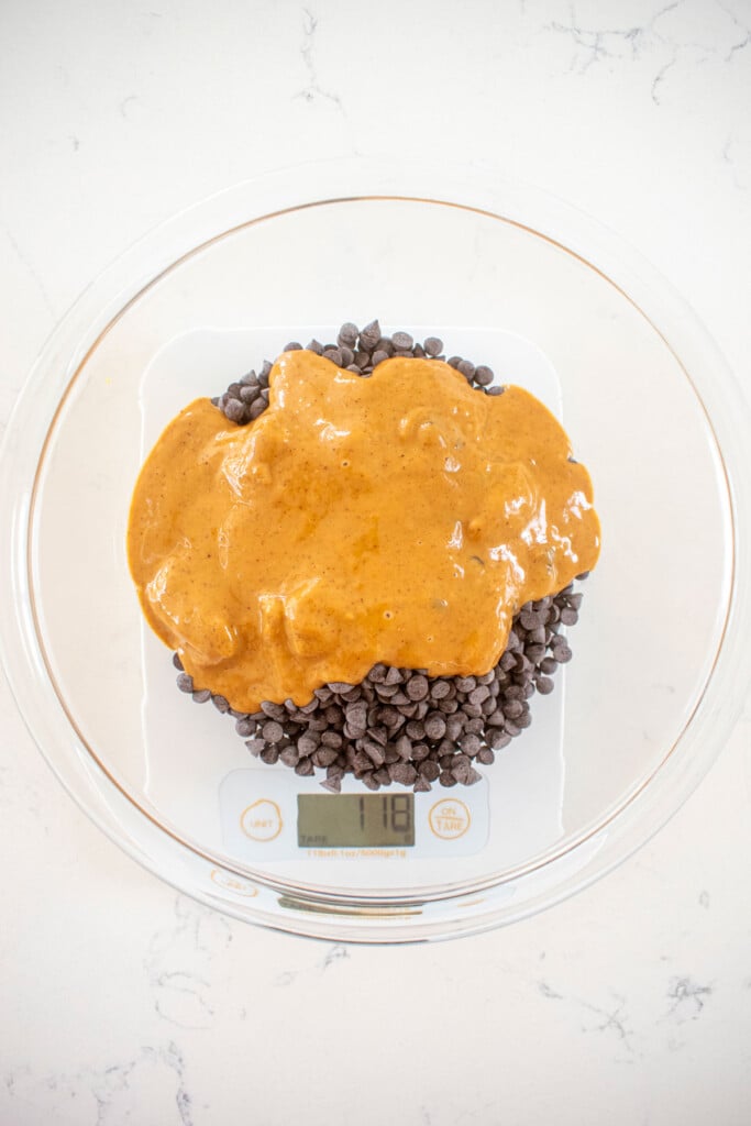 chocolate and peanut butter in a glass mixing bowl on a scale on a white marble counter.