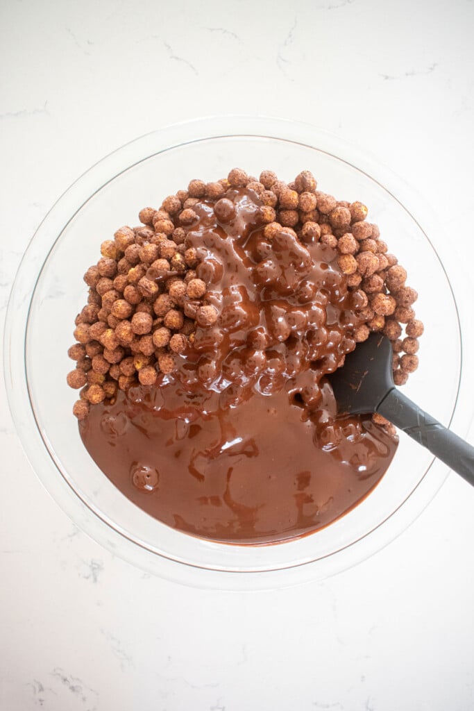 puppy chow being stirred with chocolate and peanut butter in a glass bowl on a white marble counter.