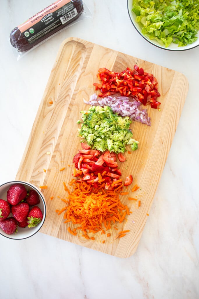 chopped veggies on a wood cutting board with a bowl of lettuce and strawberries on a marble counter.
