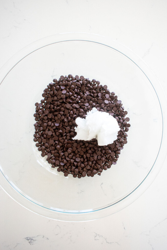 mini dark chocolate chips and coconut oil in a glass mixing bowl on a white counter.
