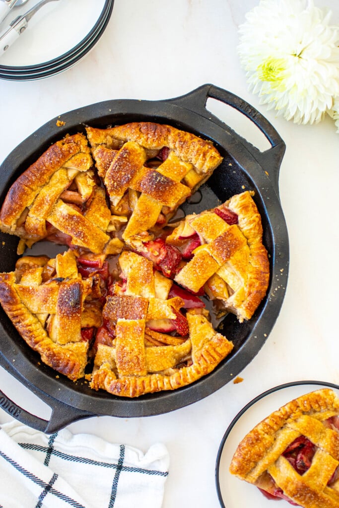 apple cinnamon strawberry pie in a cast iron pie pan cut into slices on a white marble table with fresh flowers.