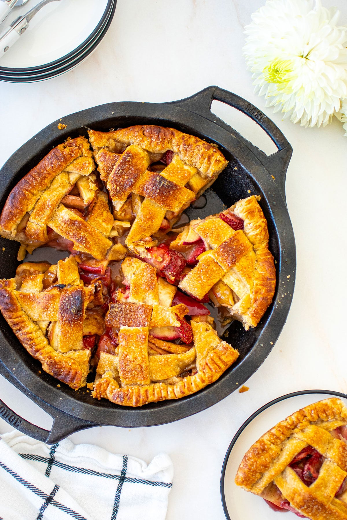 apple cinnamon strawberry pie in a cast iron pie pan cut into slices on a white marble table with fresh flowers.